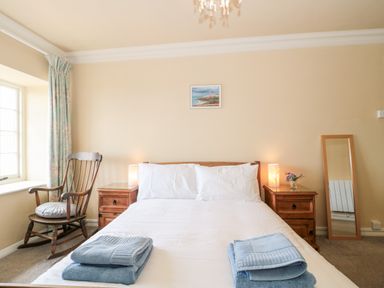 A bedroom with a bed with white bedding and folded blue towels a wooden rocking chair two wooden bedside tables lamps a standing mirror and a window with curtains at The Pound in Porlock
