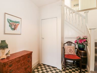 A hallway with a wooden chest of drawers a green lamp a framed bull painting a wooden chair with a cushion a vase with flowers and a staircase at The Pound in Porlock