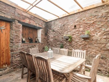 An outdoor covered dining area with a wooden table and eight wooden chairs against a stone wall at The Pound in Porlock
