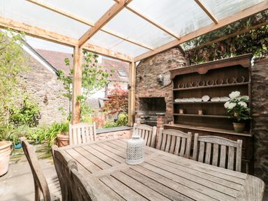 An outdoor patio with a wooden table and chairs a stone wall a wooden shelf with decorations and plants and a transparent roof at The Pound in Porlock
