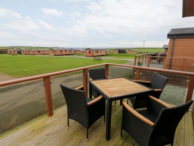 An outdoor deck with a wooden table and four black chairs overlooking a grassy area and several cabins at Reef Retreat in Broad Haven