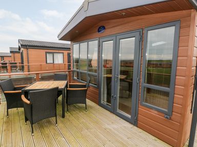 An outdoor wooden deck with a table and six chairs outside a cabin with glass doors and windows at Reef Retreat in Broad Haven
