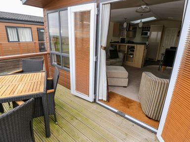 A wooden deck with a table and chairs outside a living area with armchairs and a kitchen at Reef Retreat in Broad Haven