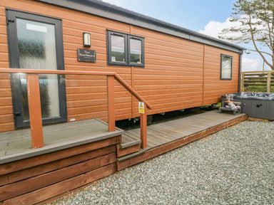 The exterior of a wooden cabin with a door window railing deck and hot tub on gravel at Rimini Retreat