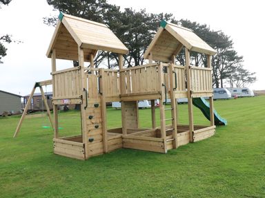 A wooden outdoor play structure with two towers connected by a bridge a slide a swing and a climbing wall on grass at Seahorse Retreat in Little Haven near Broad Haven
