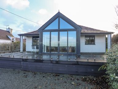 A house with large glass doors and windows facing a wooden deck with glass railing at Haulfryn in Moelfre