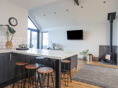 A kitchen area with a marble countertop island and wooden stools next to a living room with a wall-mounted TV and a wood stove at Haulfryn in Moelfre