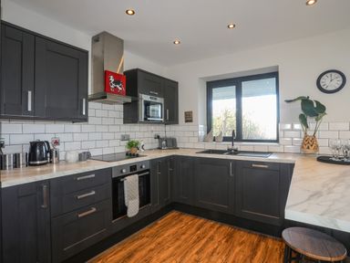A kitchen with dark cabinets a stovetop oven and microwave a window above the sink and a clock on the wall at Haulfryn in Moelfre