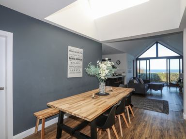 A dining area with a wooden table and black chairs next to a living room with a sofa and large glass windows showing a sea view at Haulfryn in Moelfre
