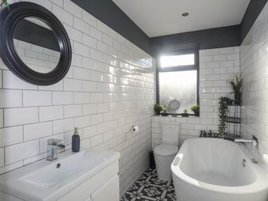 A bathroom with a white bathtub toilet and sink with soap dispenser and a round mirror on tiled walls at Haulfryn in Moelfre