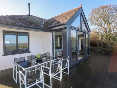 An outdoor deck with a glass dining table and chairs next to a house with large glass windows at Haulfryn in Moelfre