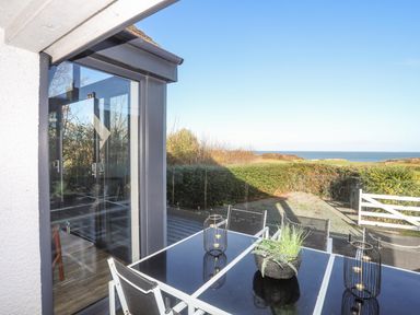 A glass outdoor dining table with chairs on a patio overlooking a hedge and a distant sea view at Haulfryn in Moelfre
