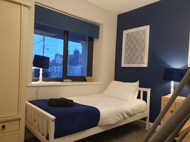A bedroom with a single bed blue blanket a window white lamp and a framed artwork on a blue wall at Haulfryn Moelfre