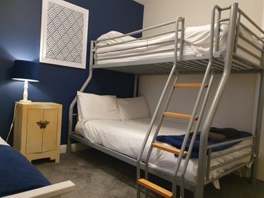 A bedroom with a metal bunk bed a wooden nightstand with a lamp and a framed geometric artwork on a blue wall at Haulfryn in Moelfre