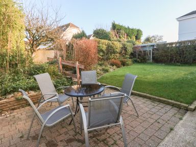 An outdoor patio area with a round glass table and five chairs next to a garden with grass and bushes at 30 Long Shepherds Drive in Newton near The Mumbles