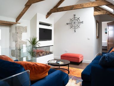 A living room with blue sofas orange cushions a black coffee table a small red ottoman a wall-mounted television a glass-enclosed fireplace a plant wooden beams and a compass wall decoration at The Rum House in Porthallow