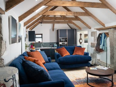 A living room with blue sofas and orange cushions with a round wooden coffee table and an open plan kitchen under wooden ceiling beams at The Rum House in Porthallow