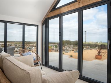 A sunroom with beige sofa and wicker chair with pillows next to large glass doors opening to a patio at The Rum House in Porthallow