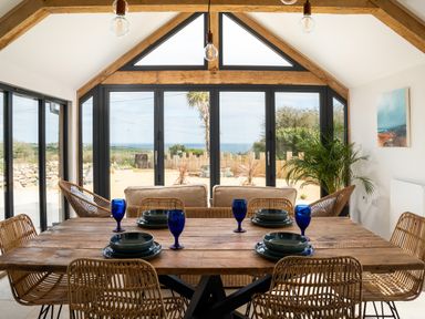 A dining area with a wooden table set with blue glasses and bowls surrounded by wicker chairs and a seating area by large windows showing a garden view at The Rum House in Porthallow