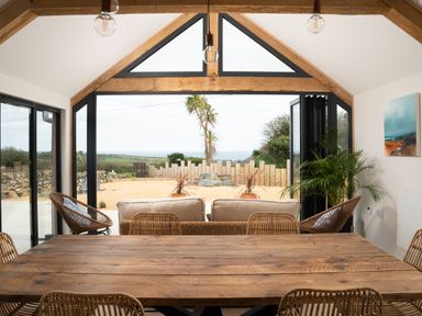 A dining area with a wooden table and wicker chairs facing a seating area with sofas and rattan chairs overlooking an outdoor space with plants at The Rum House in Porthallow