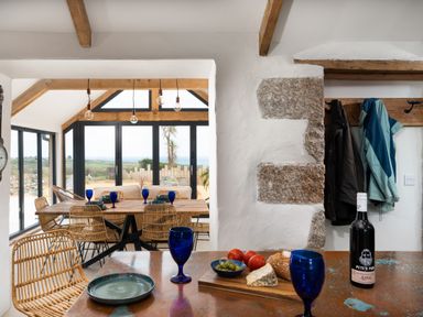 A dining area with wicker chairs wooden table blue glasses and a kitchen counter with bread cheese tomatoes olives and a bottle of wine at The Rum House in Porthallow