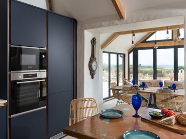 A kitchen and dining area with a wooden table set with blue glasses and plates with large windows showing a view outside at The Rum House in Porthallow