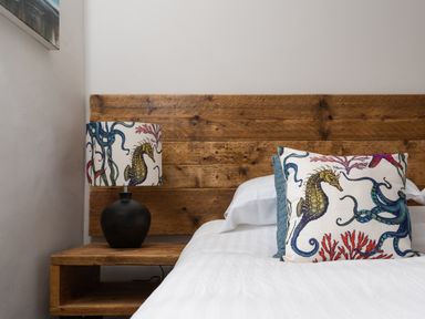 A bedroom with a wooden headboard a bedside table with a lamp and a pillow with sea creature designs at The Rum House in Porthallow