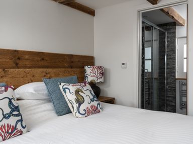 A bedroom with a wooden headboard sea-themed pillows bedside table lamp and an open door to a bathroom at The Rum House in Porthallow