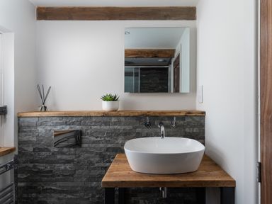 A bathroom sink on a wooden countertop with a rectangular mirror above and stone tile wall at The Rum House in Porthallow