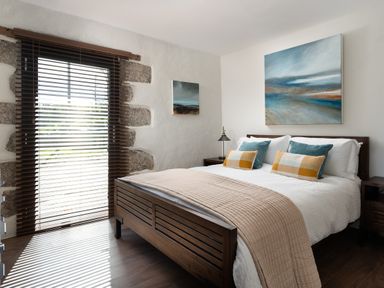 A bedroom with a wooden bed and nightstands a large window with blinds and abstract paintings on walls at The Rum House in Porthallow