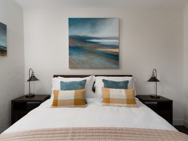 A bedroom with a bed between two bedside tables with lamps and a painting above the bed at The Rum House in Porthallow