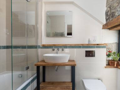 A bathroom with a wooden sink stand a white sink mirror glass shower screen toilet and a plant on a windowsill at The Rum House in Porthallow