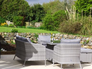 An outdoor patio with wicker furniture a glass table a bottle of wine and wine glasses with a stone wall and garden in the background at The Rum House in Porthallow