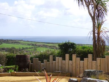 An outdoor garden area with a wooden fence a barrel and a tree overlooking green fields and the sea at The Rum House in Porthallow
