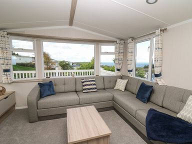 A living room with a gray sectional sofa and cushions with a wooden coffee table and windows showing outdoor greenery at Gwent 74 in Borth