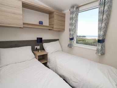 A bedroom with two single beds a bedside table with a lamp and a window with curtains overlooking a landscape at Gwent 74 in Borth