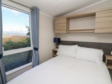 A bedroom with a bed white pillows a bedside table with a lamp and a window with blue curtains at Gwent 74 in Borth