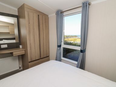 A bedroom with a white bedspread a wooden wardrobe and a window with blue curtains showing a view outside at Gwent 74 in Borth