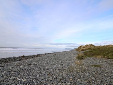 A pebble beach with grass-covered dunes and the sea under a partly cloudy sky at Gwent 74 in Borth