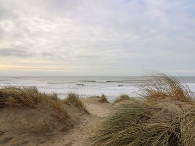 A sandy beach with grass-covered dunes and waves in the ocean under a cloudy sky at Gwent 74 in Borth