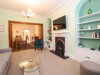 A living room with a fireplace a wall mounted TV a blue chair a glass coffee table and bookshelves opening to a dining area at Blencathra in Keswick