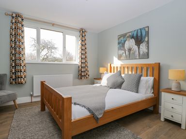 A bedroom with a wooden bed frame grey pillows two bedside tables with lamps and windows with patterned curtains at Birch House in Corsham
