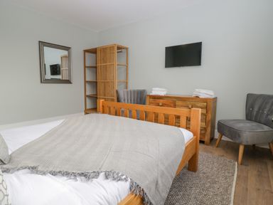 A bedroom with a wooden bed a gray blanket two gray chairs a wooden dresser a wooden shelf a mirror and a wall-mounted TV at Birch House in Corsham
