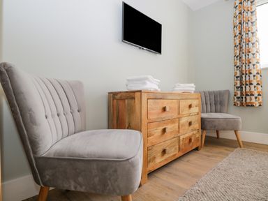A room with two grey chairs a wooden dresser with folded white towels and a wall mounted TV at Birch House in Corsham