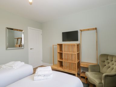 A bedroom with two beds, folded white towels on each, a wooden cabinet, a mounted television, a green armchair, a full-length mirror, and a white door at Birch House in Corsham