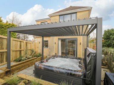 A backyard with a hot tub under a pergola and a stone house with sliding glass doors at Birch House in Corsham