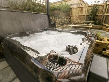 An outdoor hot tub with bubbling water near a wooden picnic table and fenced garden at Birch House in Corsham