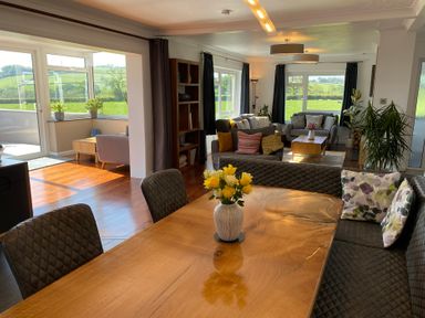 A dining table with a flower vase and cushioned chairs next to a living area with sofas and large windows at Ashlea in Launceston