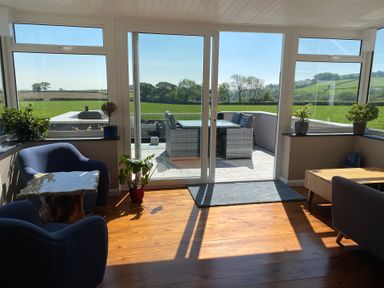 A sunlit living room with blue chairs plants and a wooden table overlooking a patio with outdoor furniture and green fields at Ashlea Launceston