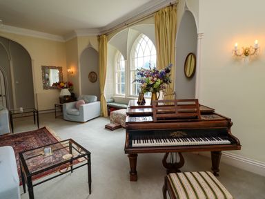 A room with a piano, armchairs, glass tables, large window with curtains and a floral arrangement at Craster Tower Penthouse in Craster